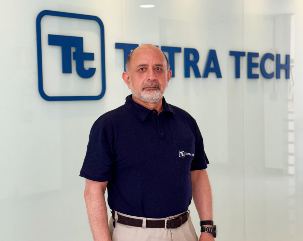 Homem branco, calvo, com barba branca. Ele veste uniforme azul com inscrições da Tetra Tech no lado esquerdo. Ao fundo, há um letreiro com o logotipo da Tetra Tech.